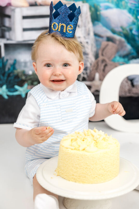 Baby boy in "one" crown sitting next to first birthday cake