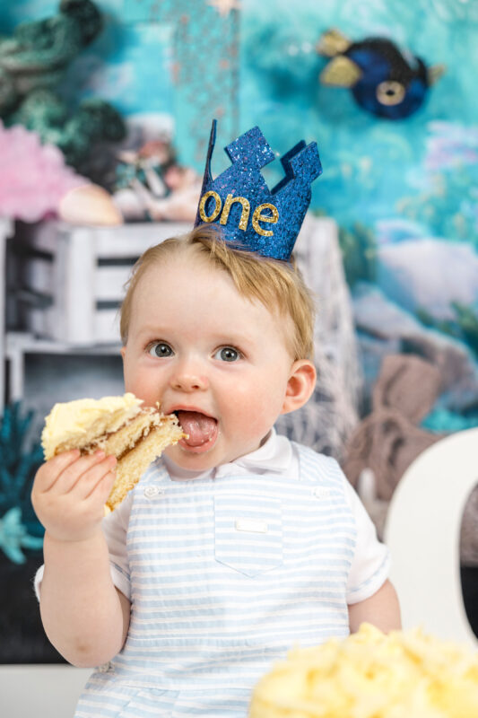Happy baby boy eating first birthday cake with "one" hat