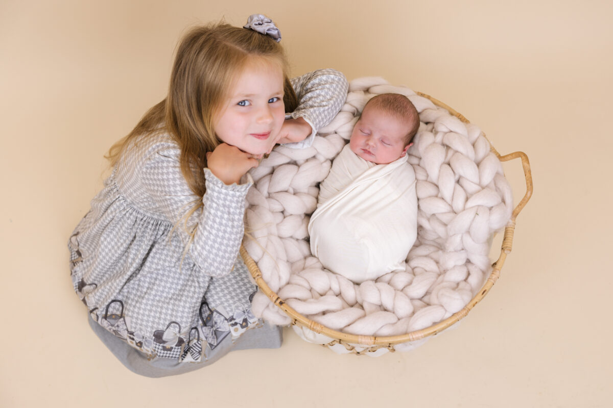 Big sister looking at her sleeping newborn sibling in a basket with a chunky knit blanket during a sibling photoshoot at Little Miracles Photography Studio.