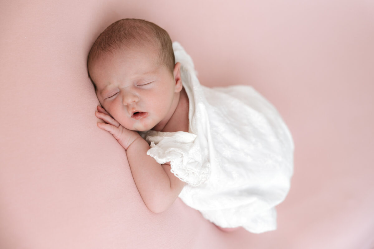 Sleeping newborn baby girl wrapped in white, lying on a pink blanket during a photoshoot with Little Miracles Photography Studio.