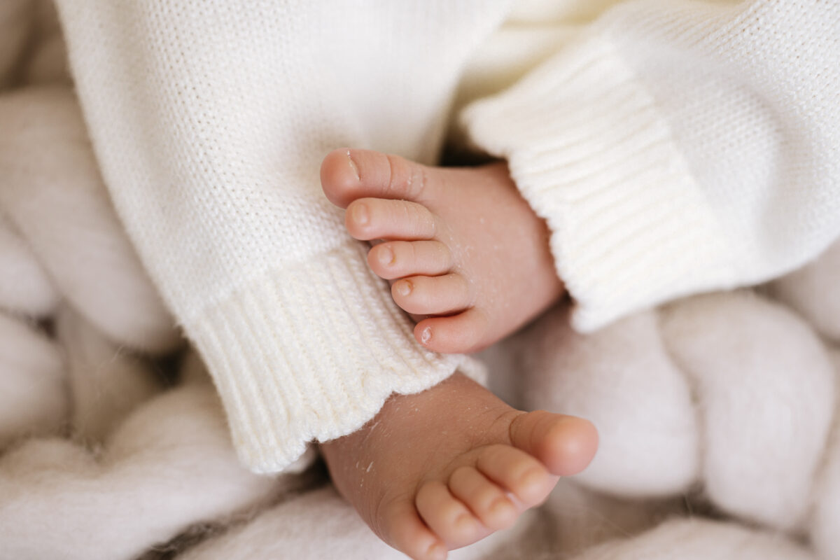 Close-up of tiny newborn baby feet wearing white knitted trousers, resting on a soft white blanket during a photoshoot.