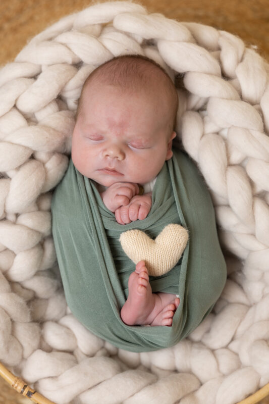 Peaceful newborn baby boy awrapped in green fabric, posed on chunky wool blanket during a studio newborn photography session in Lanarkshire at Little Miracles Photography Studio.