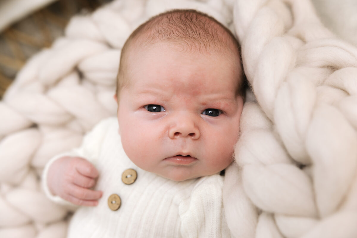 Newborn baby gazing at camera while wrapped in a soft ivory chunky knit blanket, wearing cream ribbed outfit with wooden buttons in a newborn photography session