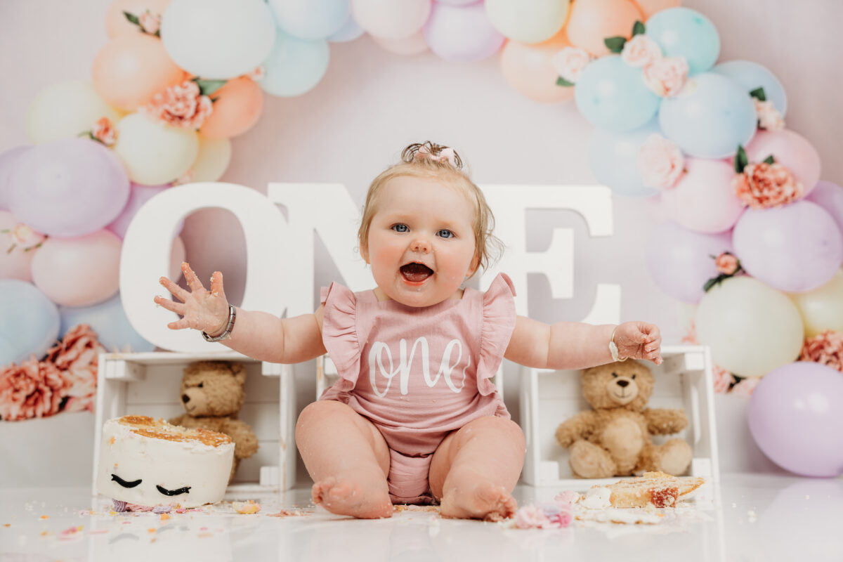 Happy baby celebrating a first birthday with a cake smash photoshoot, surrounded by balloons and decor at Little Miracles Photography Studio.