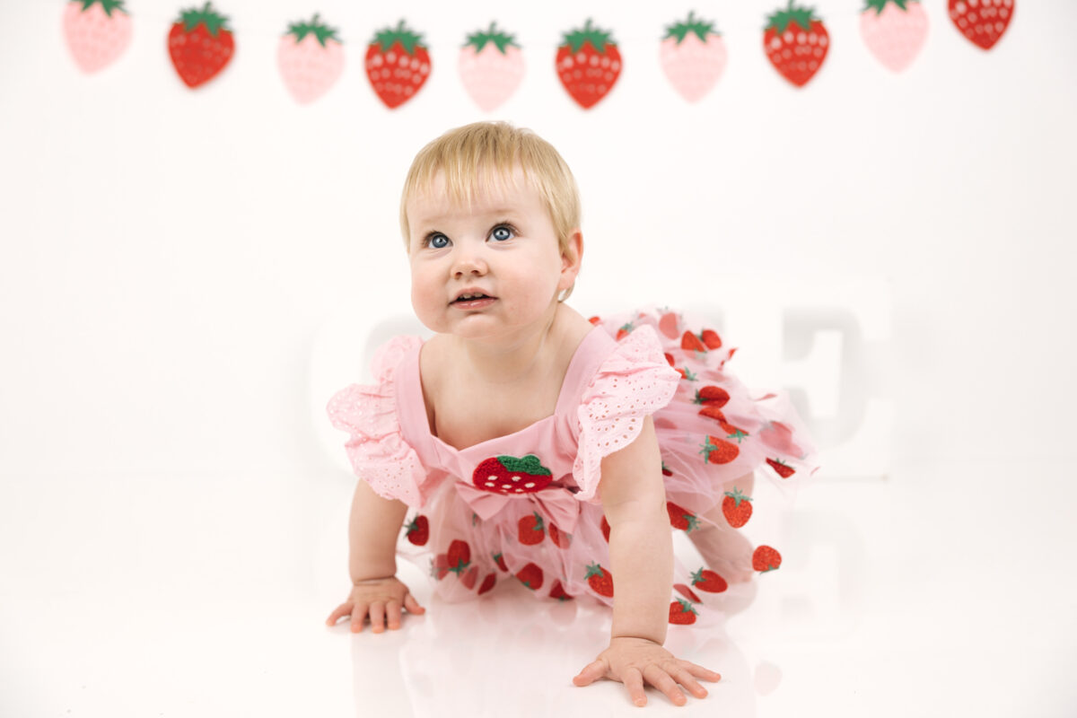 Smiling baby girl celebrating her first birthday with pink and red strawberry bunting.