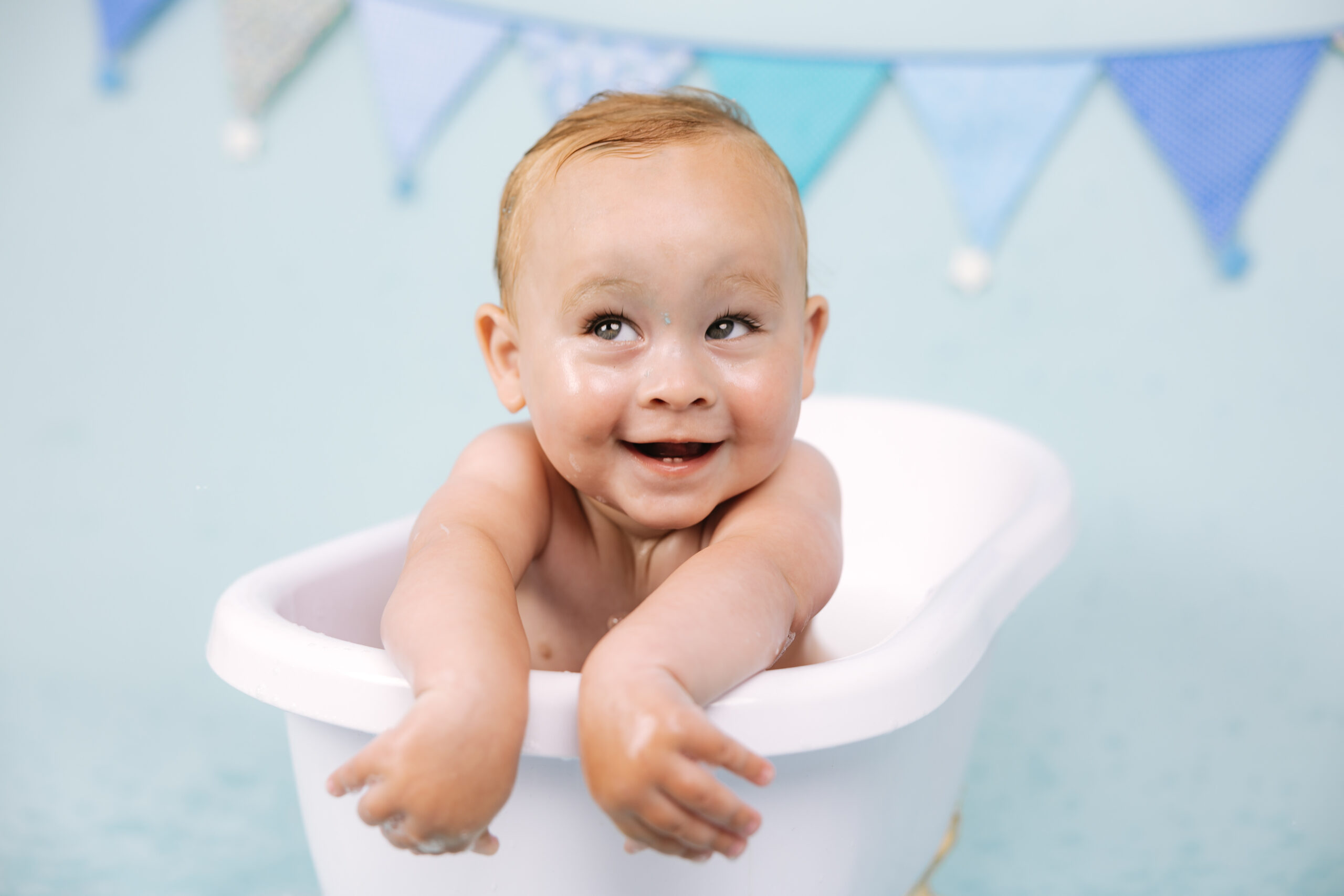 Baby boy enjoying a splash bath after his first birthday cake smash against a soft blue studio backdrop.