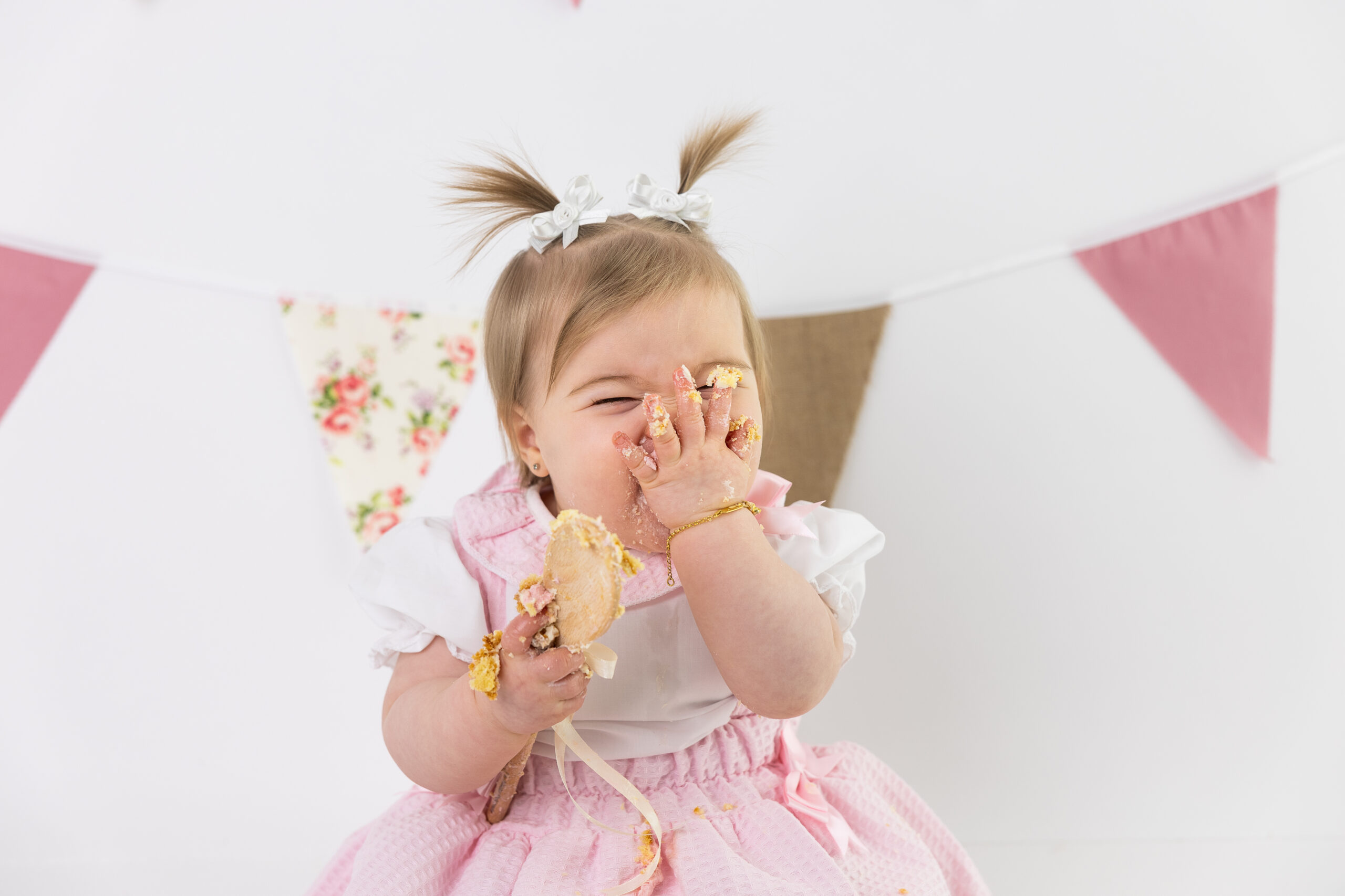 Smiling baby girl eating birthday cake against a white studio backdrop.