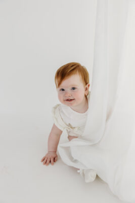 Baby girl peeking behind white curtain during first birthday photoshoot