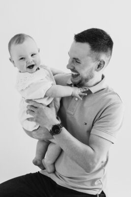 Smiling dad photographed with baby girl against a neutral studio backdrop.