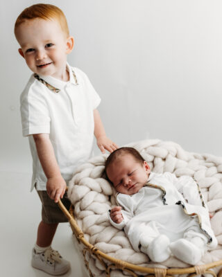 Older brother standing beside newborn baby in wicker basket.