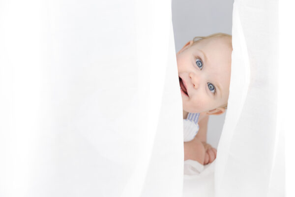 Baby boy peeking around a white curtain.