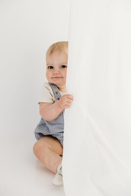Baby boy peeking from behind a white curtain.