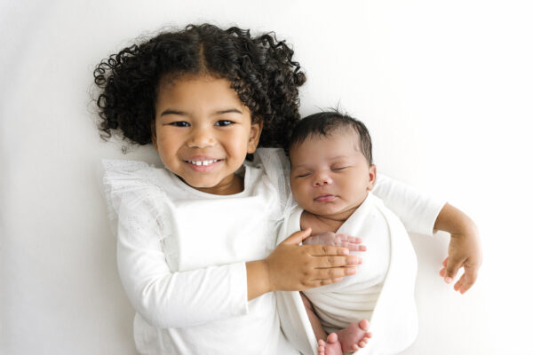 Smiling older sibling holding newborn baby on white background.